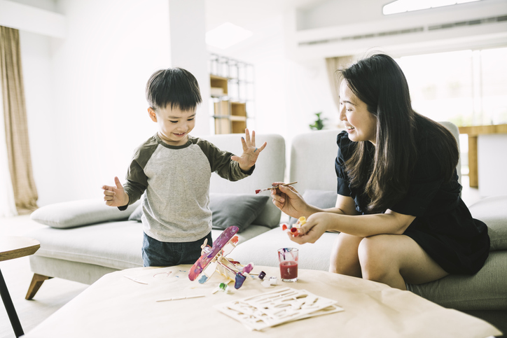 Smiling mother painting model airplane for cute son. Female parent and child are enjoying the time together in living room.